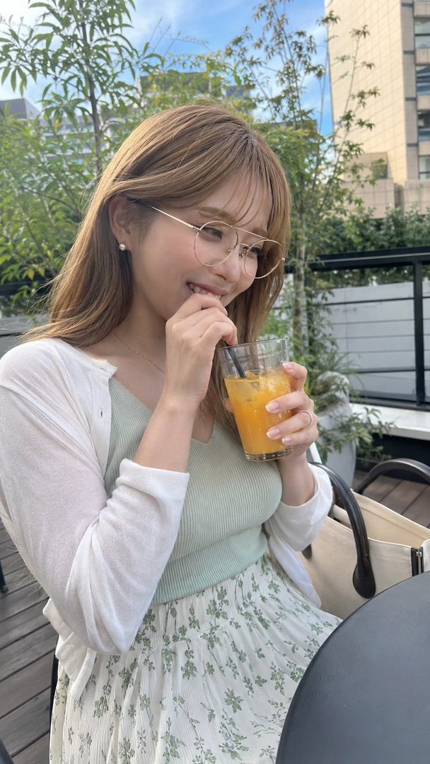 A young woman with long brown hair and glasses, wearing a light green top and a floral skirt, drinks an orange beverage on a sunny outdoor terrace.