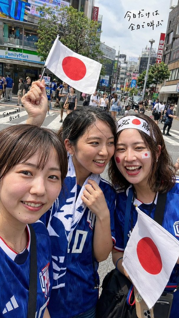 Three young women in blue soccer jerseys and Japanese flag face paint take a joyful selfie on a city street, holding small Japanese flags.