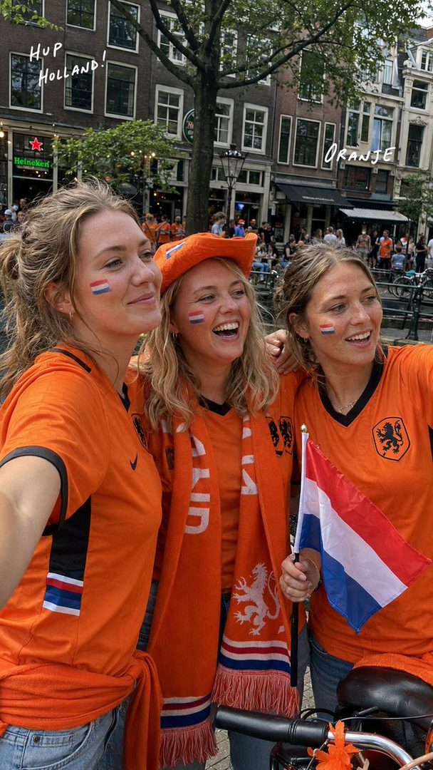 Three women in orange Netherlands football jerseys, one wearing a matching hat and scarf, smiling and posing for a selfie on a city street.