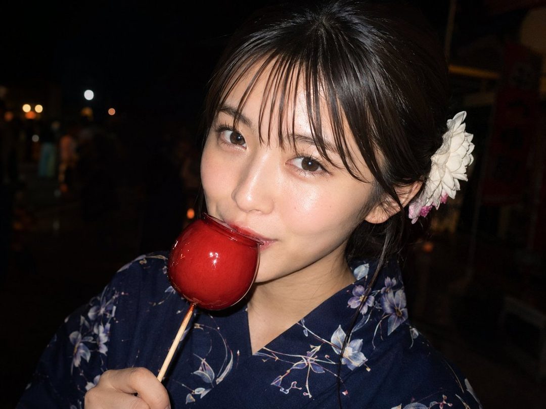 A young East Asian woman in a dark blue floral yukata eats a red candy apple at night, with a white flower in her hair.