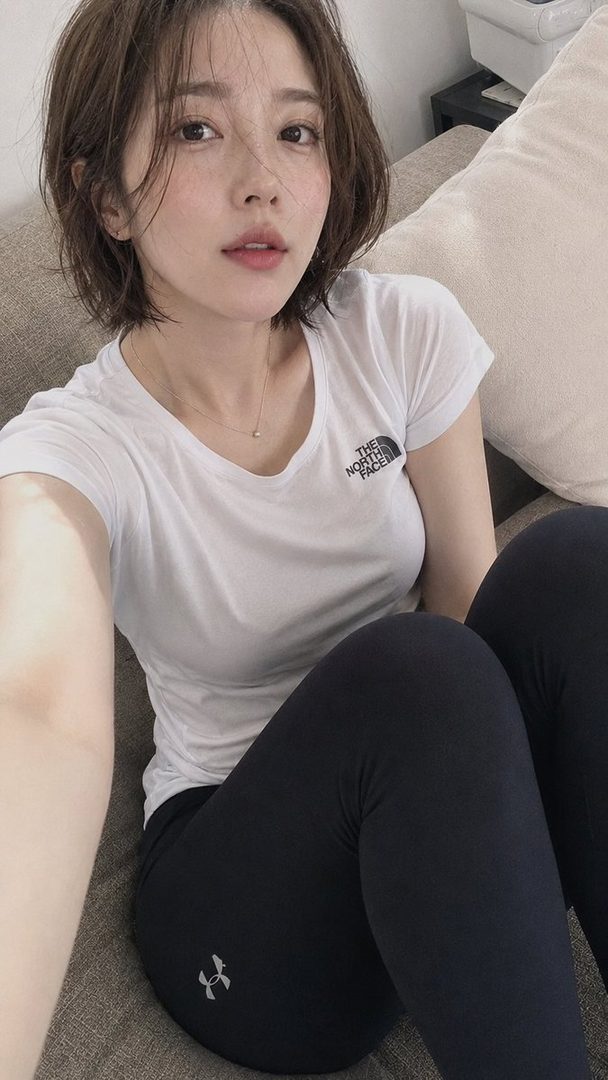 A young East Asian woman with short brown hair takes a selfie indoors, wearing a white t-shirt and black leggings.