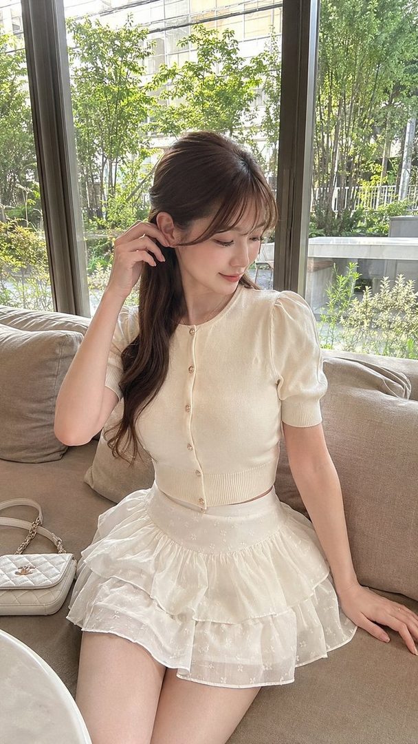 A young East Asian woman with long brown hair, wearing a cream-colored puff-sleeve cardigan and a ruffled white skirt, sits indoors on a sofa, posing elegantly.