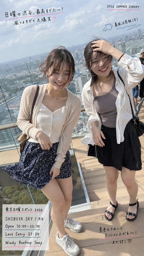 Two young women are laughing on a windy rooftop overlooking a city, dressed in casual summer outfits.