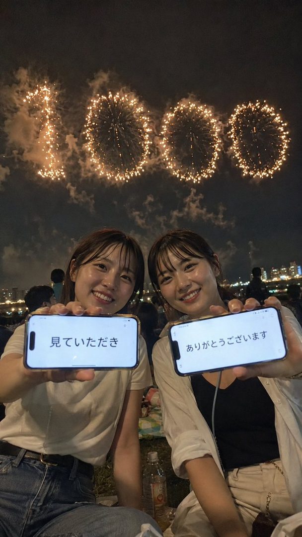 Two smiling women hold up smartphones displaying Japanese text, with fireworks shaped like the number 1000 in the night sky behind them.