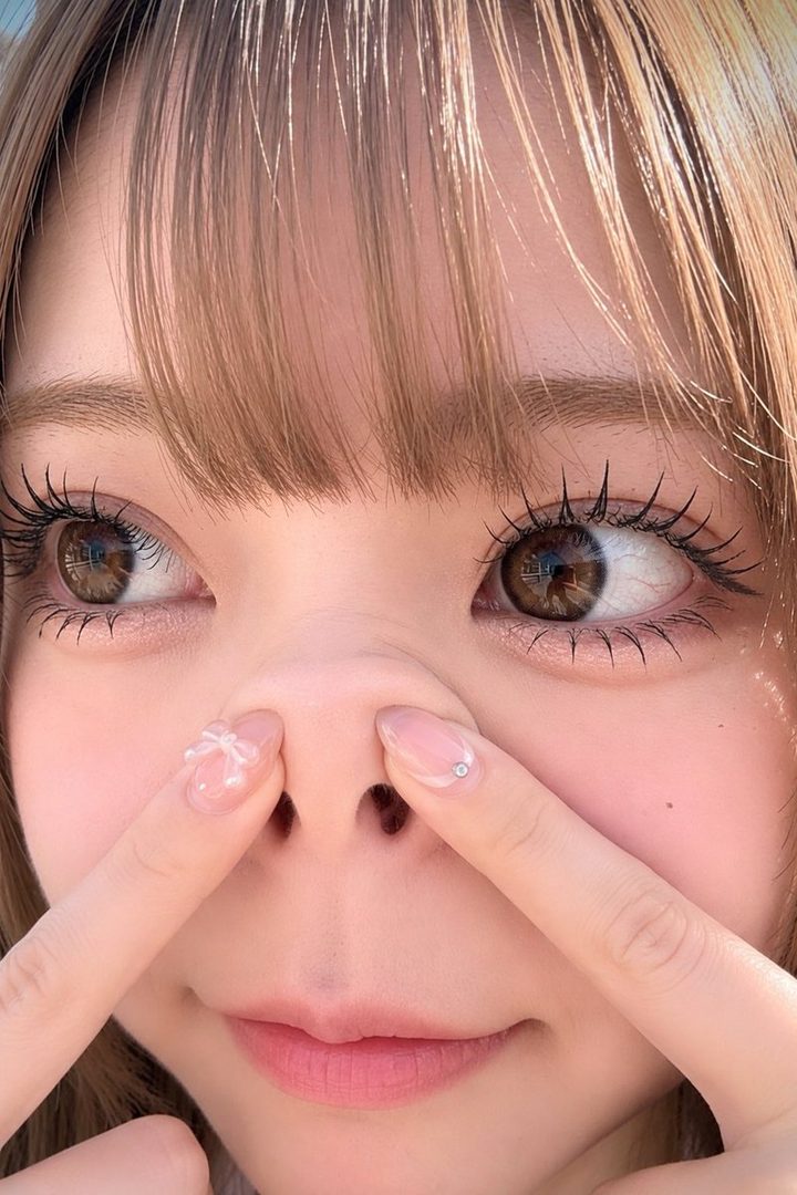 Close-up portrait of a young Japanese woman with blonde hair and bangs, looking to the side with a playful expression, her fingers touching her nose.
