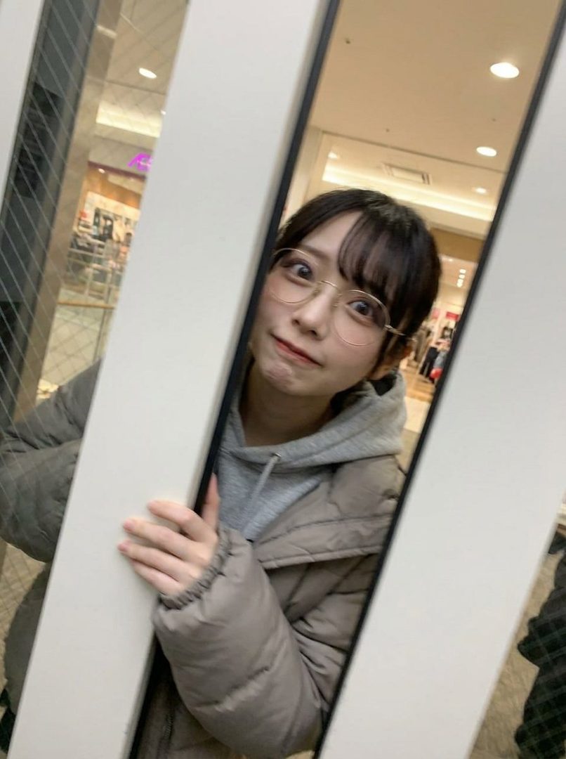 A young East Asian woman with bangs and glasses, wearing a grey hoodie and a puffer jacket, takes a selfie in a shopping mall elevator.
