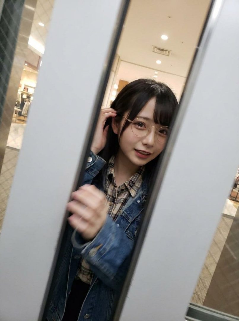A young woman with black hair and bangs, wearing glasses, a plaid shirt, and a denim jacket, poses for a selfie in an elevator.