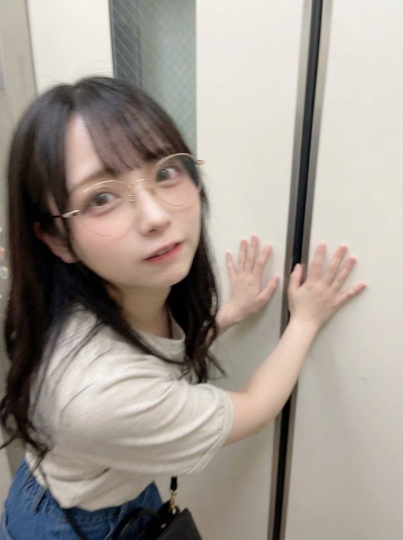 A young Japanese woman with long black hair and glasses poses for a selfie in an elevator, wearing a casual t-shirt and denim skirt.