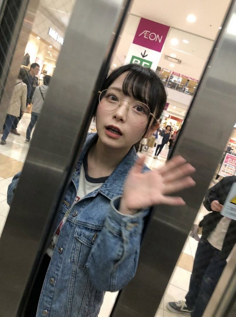 A young Japanese woman with black hair and bangs, wearing glasses and a denim jacket, waves in an elevator.