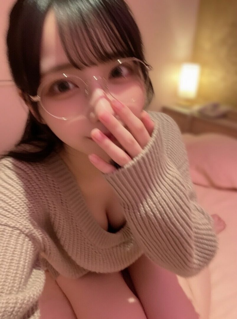 A young Japanese woman with black hair and bangs, wearing glasses and a beige sweater, poses for a selfie indoors.