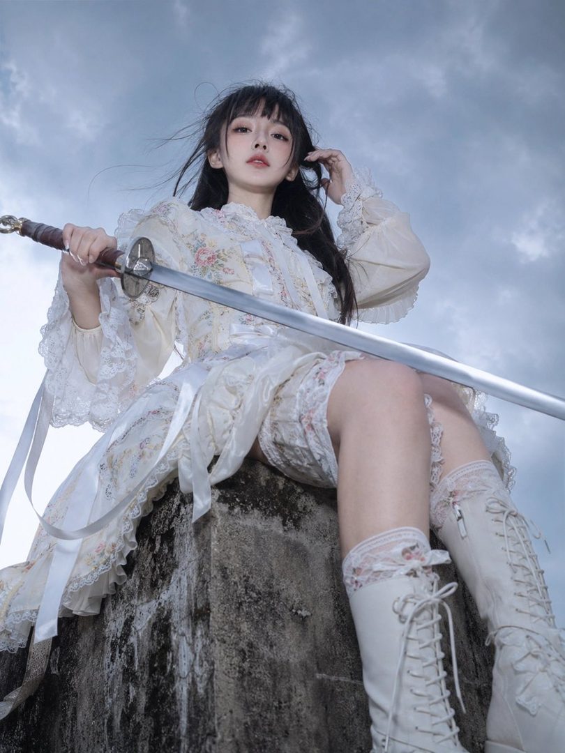 A young East Asian woman in a white lolita-inspired fantasy dress sits on a stone pillar holding a sword, with an overcast sky in the background.