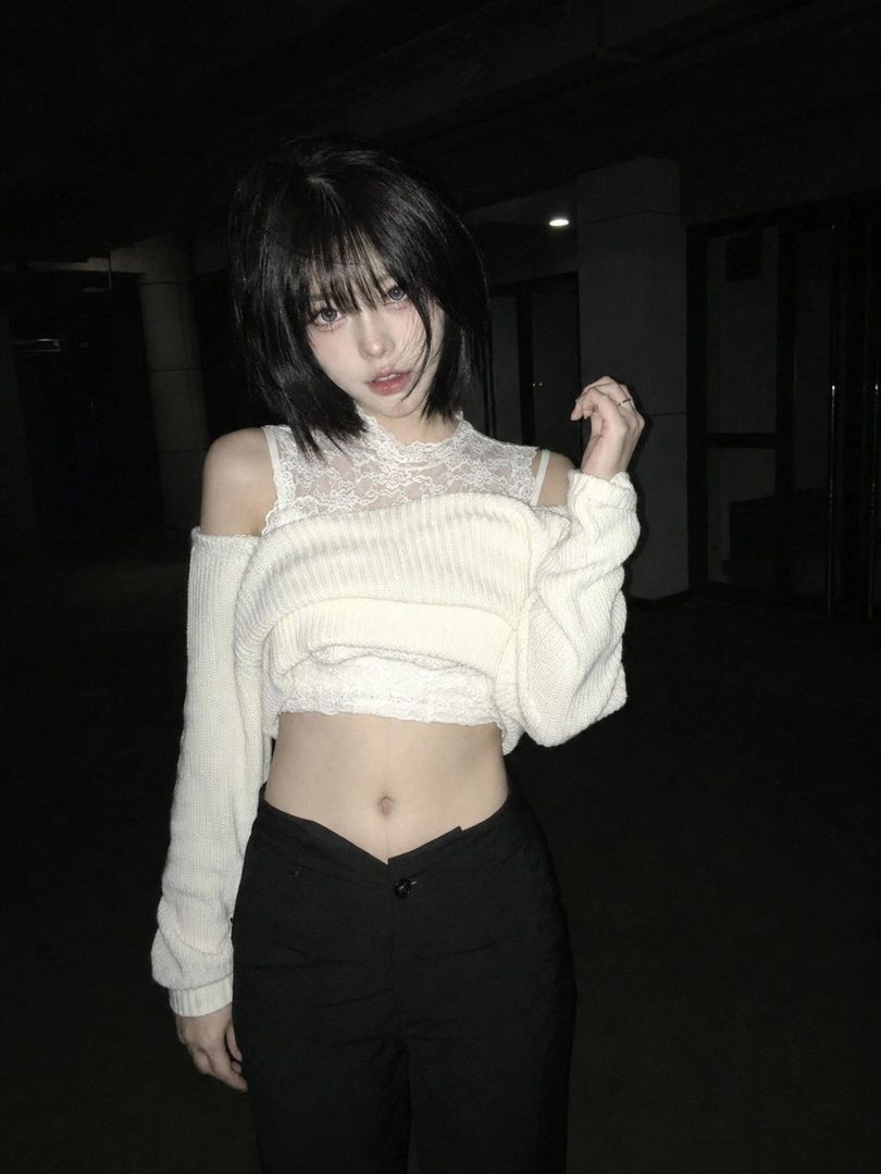 A young East Asian woman with short black hair and bangs, wearing a white lace top and an off-shoulder knit sweater, posing in a dark indoor setting with a phone flash effect.