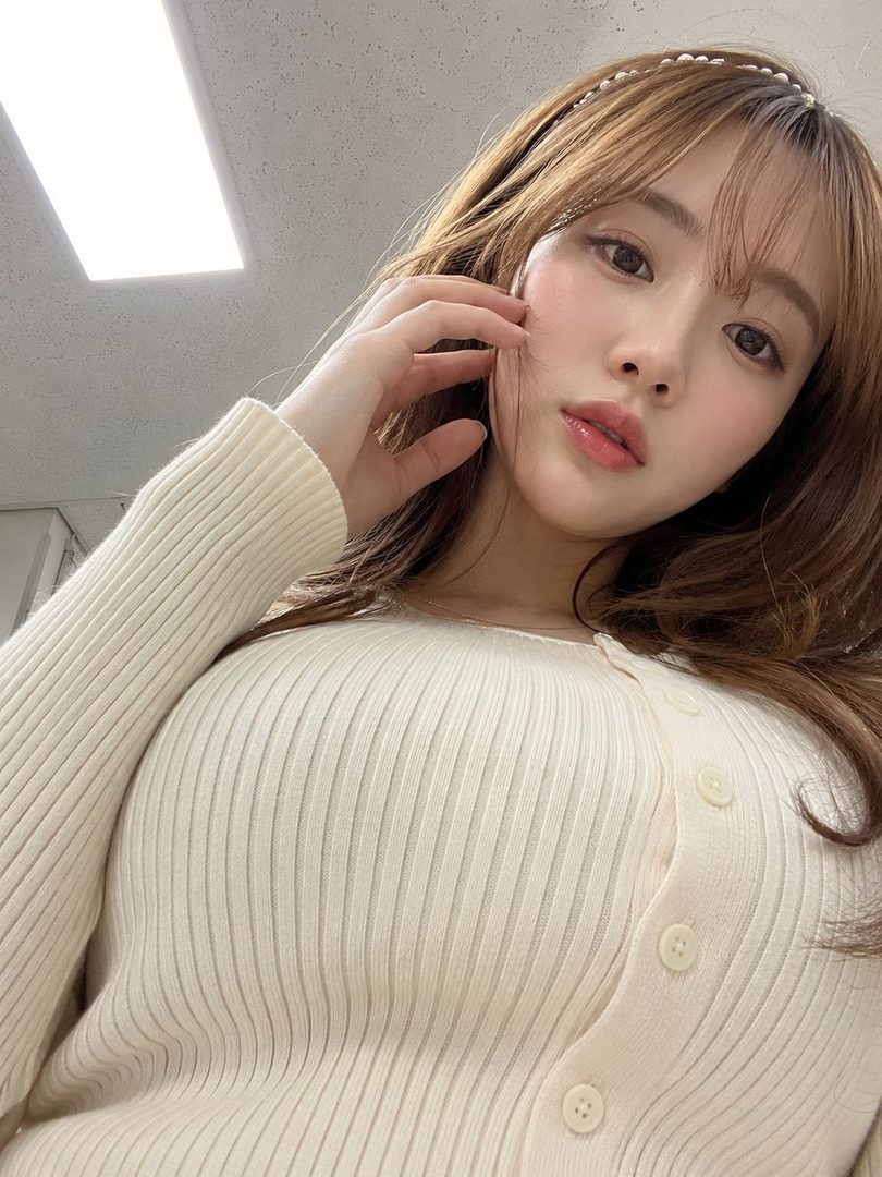 A close-up selfie of a young East Asian woman with long brown hair, wearing a cream-colored ribbed knit cardigan and a pearl headband. She is touching her face with her hand, looking at the camera with a soft expression.