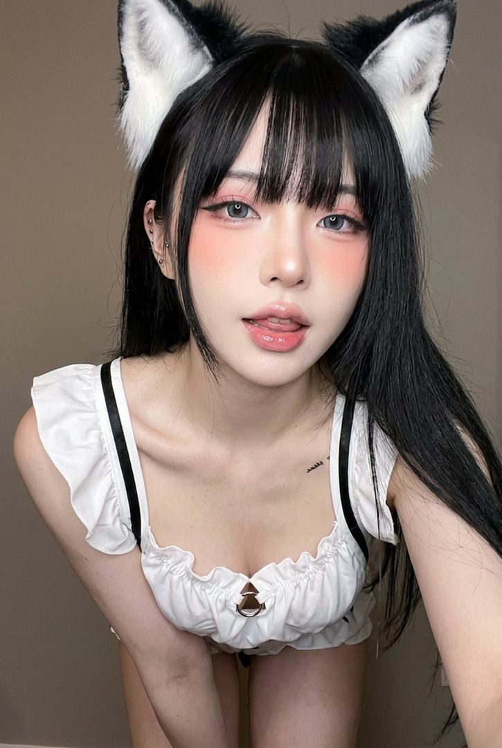 A young East Asian woman with long black hair and bangs, wearing fluffy black and white cat ears and a white ruffled top, poses for a close-up selfie with a playful expression.