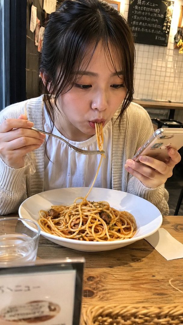 A young woman with dark hair tied up, wearing a white blouse and light cardigan, is eating a plate of spaghetti with a fork while looking at her smartphone in a casual restaurant setting.