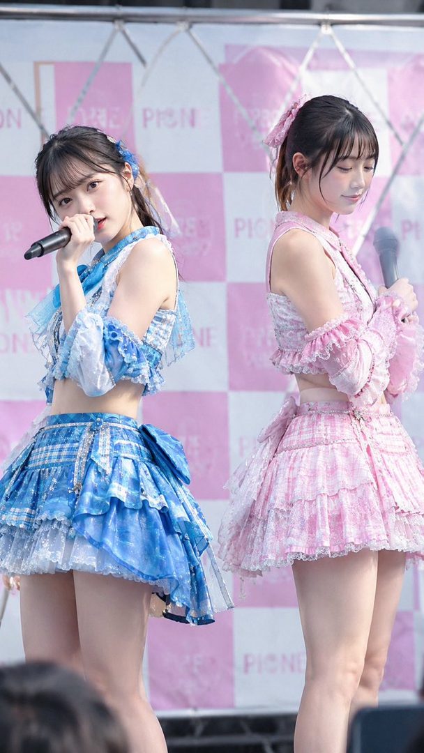 Two young women in matching blue and pink idol outfits, holding microphones on stage.