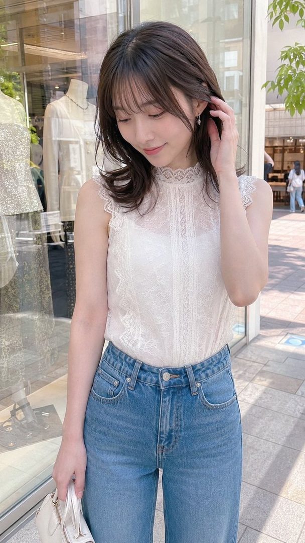 A young East Asian woman with long brown hair, wearing a white lace top and blue jeans, stands on a city street.