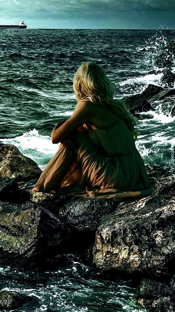 A woman with blonde hair sits on rocks by the sea, looking out at the ocean.