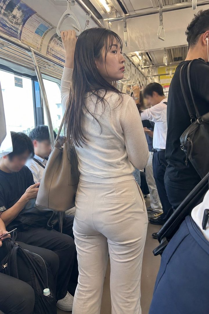 A woman with long dark hair, wearing a light sweater and beige pants, stands in a crowded subway car, holding onto a handrail.