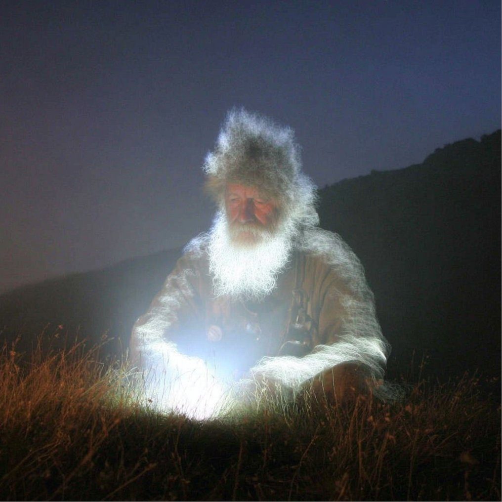 An elderly man with a long white beard and wild hair sits in a grassy field at night, illuminated by a bright light source he holds.
