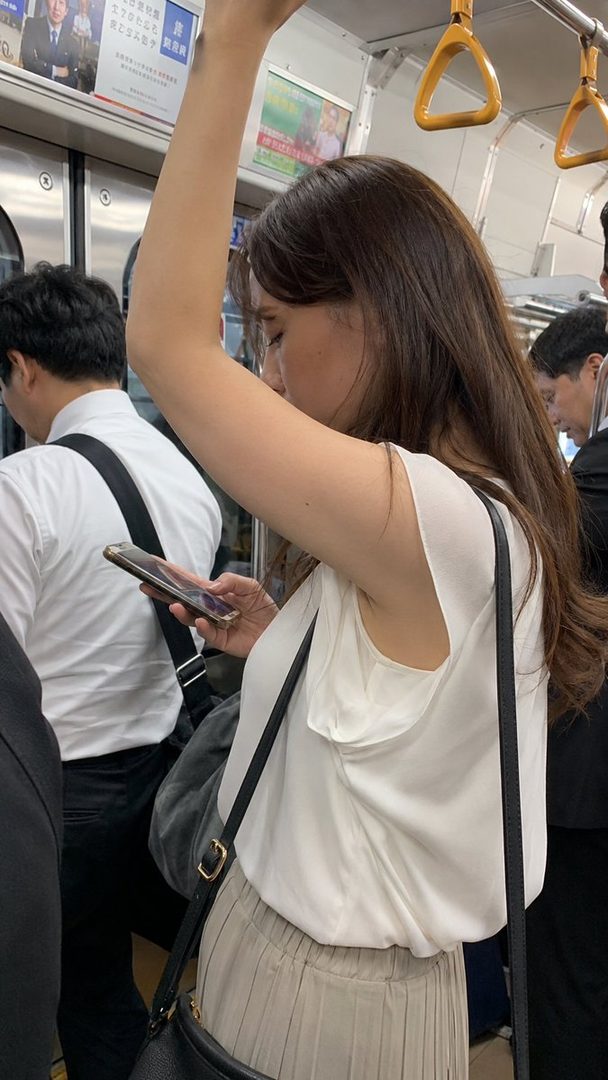 A woman with long brown hair is standing in a crowded subway train, holding onto a strap and looking at her smartphone.