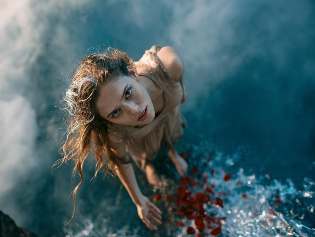 A woman with long, wavy blonde hair and striking green eyes looks up from a body of water, with red petals scattered around her.