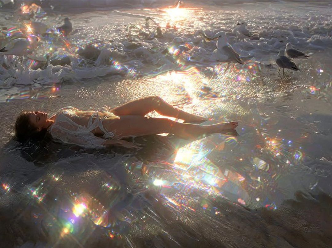 A woman in a white lace outfit lies on a beach as waves wash over her, with seagulls in the background and sparkling light effects.