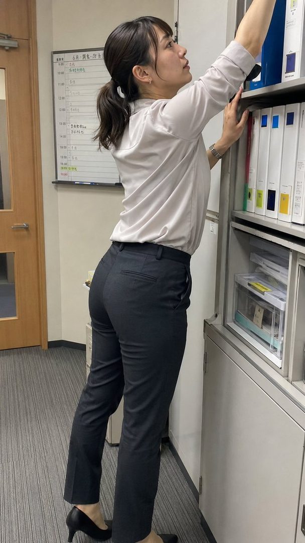 A woman in a light blouse and dark trousers reaches for files on a shelf in an office setting.