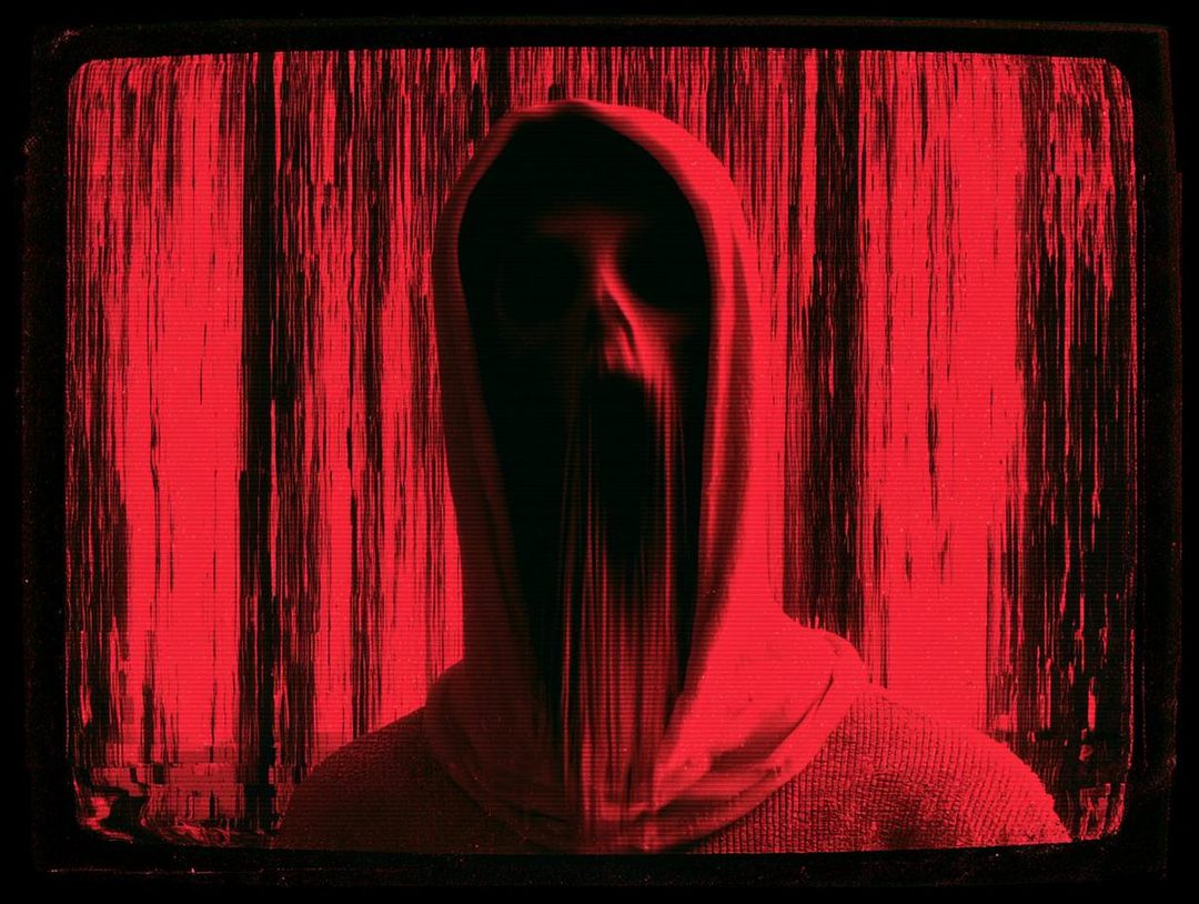 A person in a red hooded robe with distorted facial features, appearing on a glitching screen.