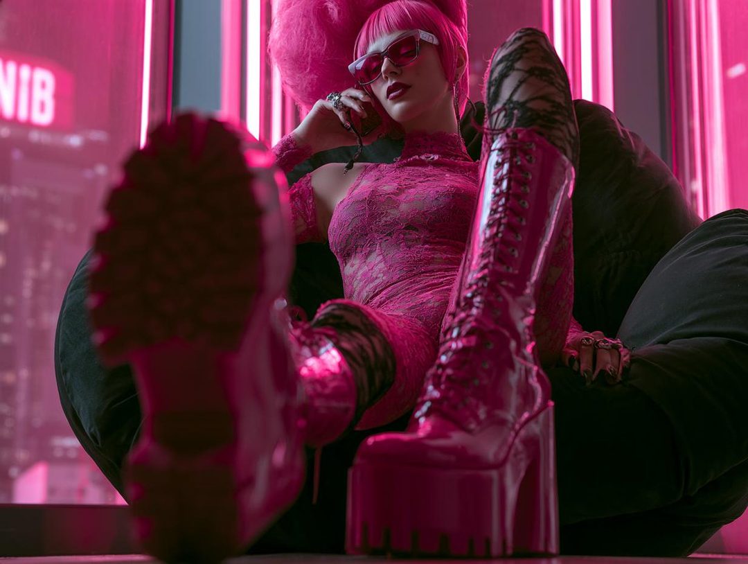 A woman with pink hair and sunglasses, wearing a pink lace dress and platform boots, poses indoors with a neon city background.