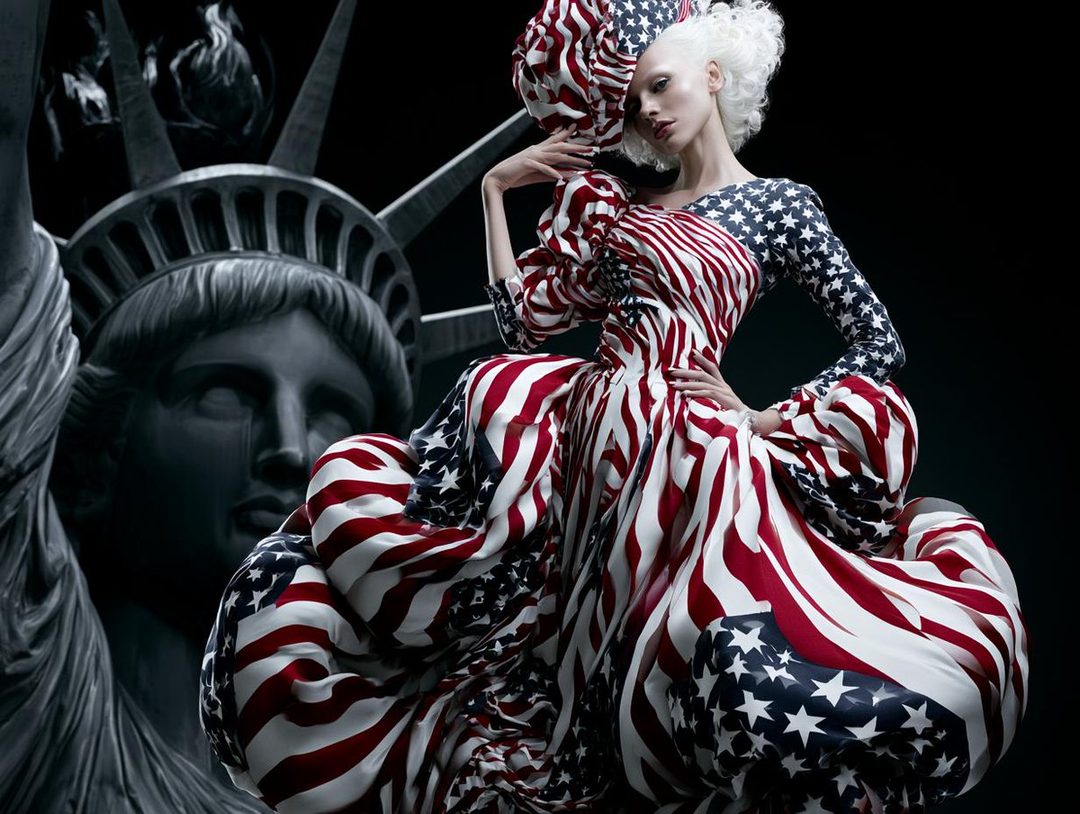 A model in a dramatic, voluminous dress patterned with the American flag poses in front of a grayscale Statue of Liberty.