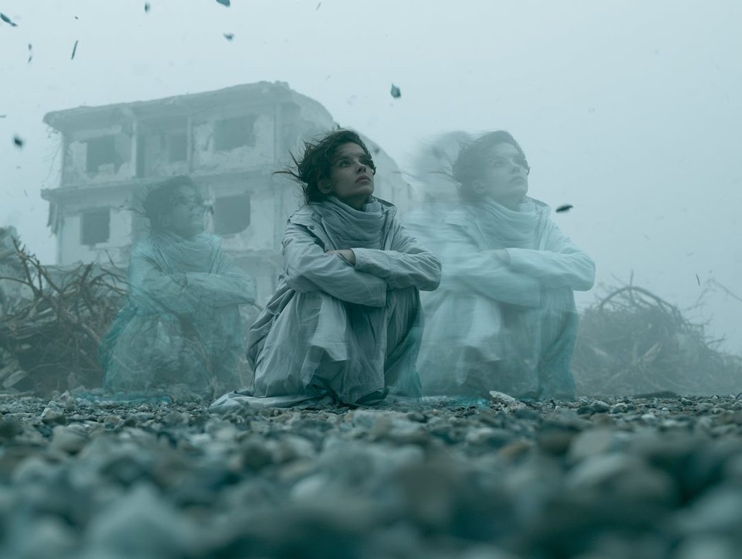 A young woman sits in a desolate, rocky landscape with a ruined building in the background, appearing as multiple ghostly figures.