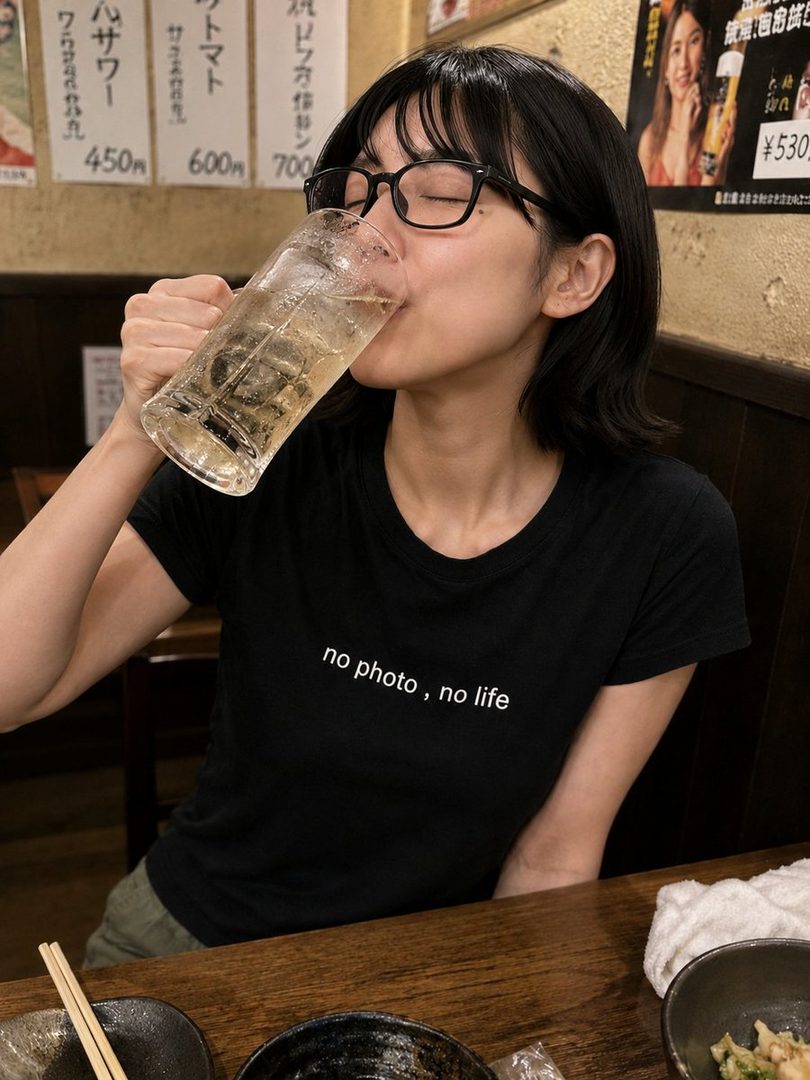 A young woman with short black hair and glasses drinks from a large glass of iced beverage in a dimly lit restaurant.