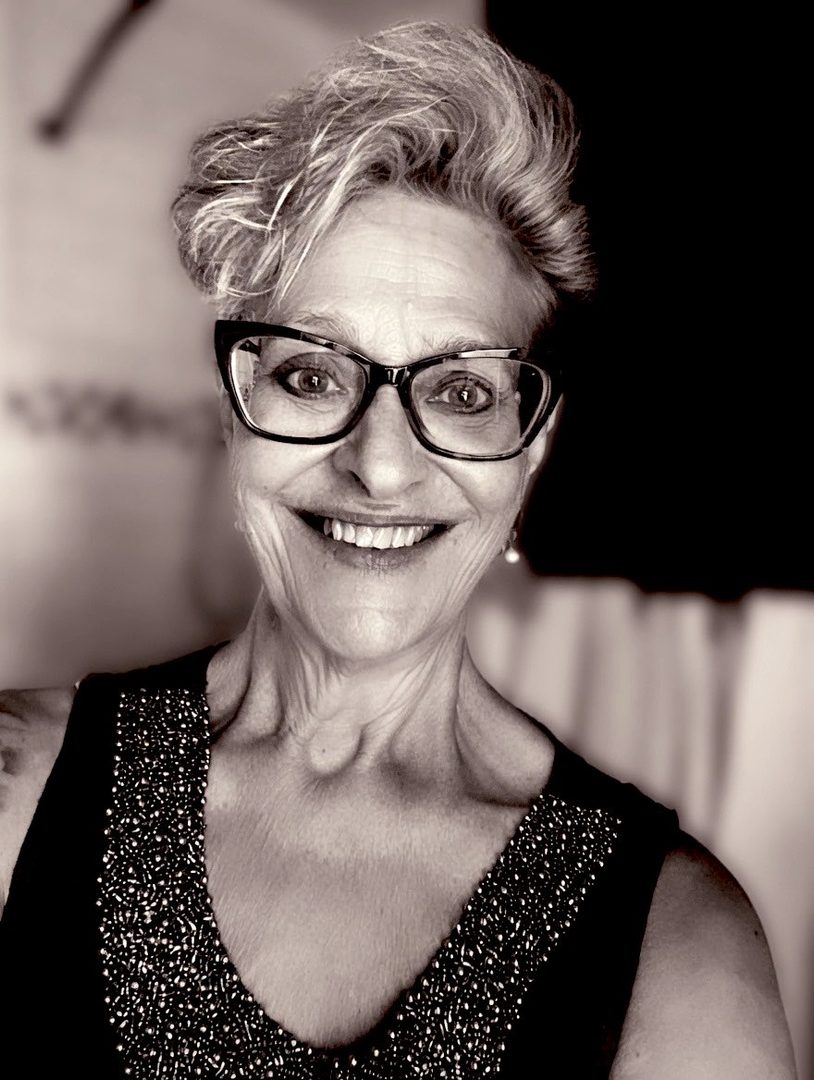 A sepia-toned portrait of an older woman with short, styled blonde hair, wearing glasses and a beaded black dress, smiling.