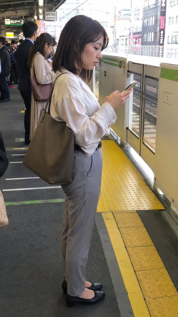 A woman with dark brown bobbed hair stands on a train station platform, looking at her smartphone. She is wearing a white blouse, grey trousers, and black flats, with a grey tote bag on her shoulder.