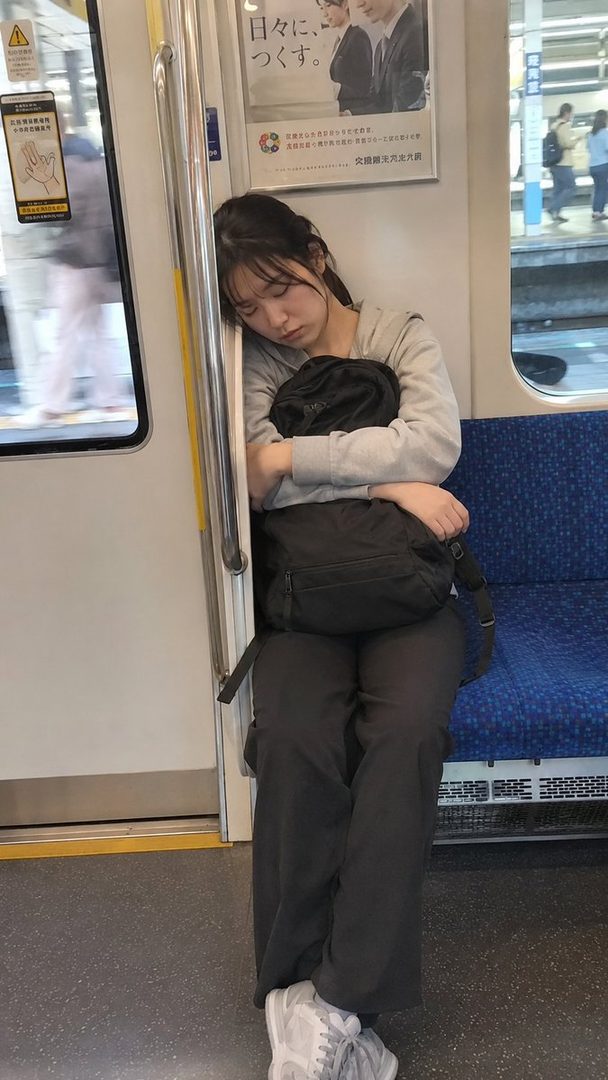 A young woman with dark hair tied in a ponytail is sleeping on a subway train, leaning against a pole and holding a black bag.
