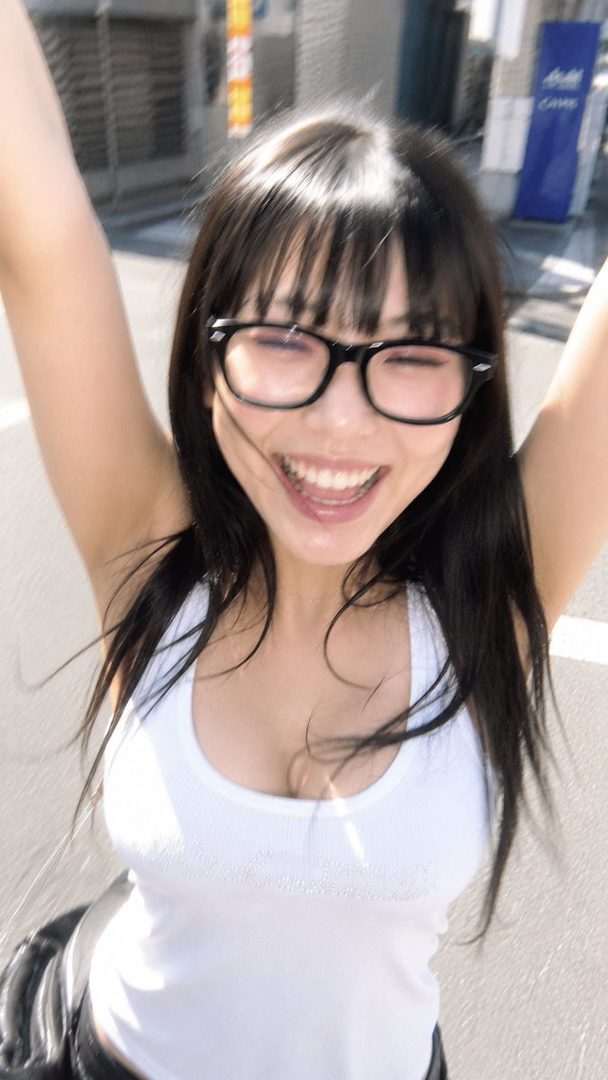 A young woman with black hair and bangs, wearing glasses and a white tank top, smiles widely with her arms raised.