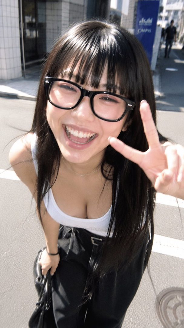 A young woman with long black hair and bangs, wearing glasses and a white tank top, smiles and makes a peace sign while posing on a city street.