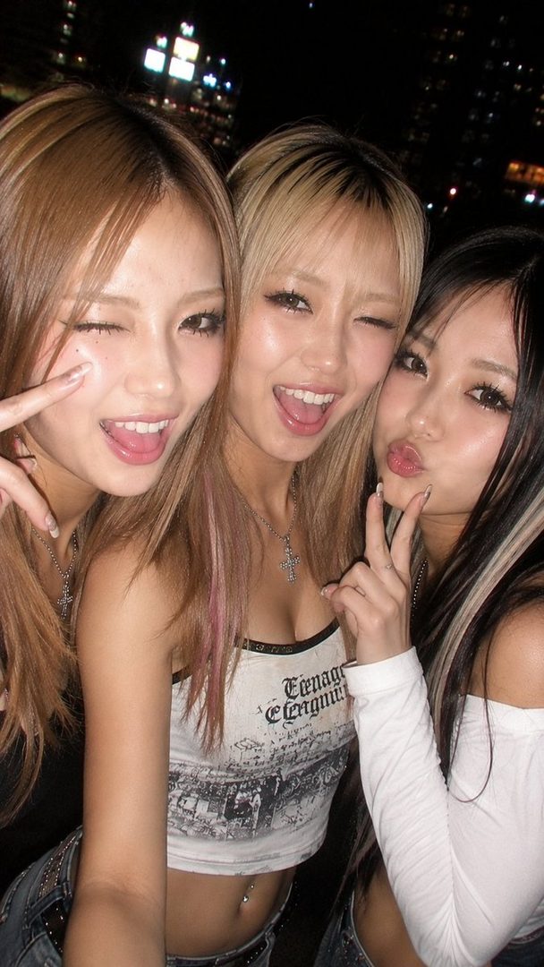 Three young East Asian women with blonde and brown hair posing for a selfie at night, winking and making peace signs.
