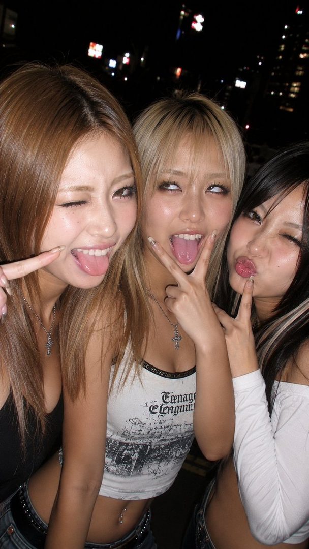 Three young East Asian women posing playfully for a selfie at night, with city lights in the background.