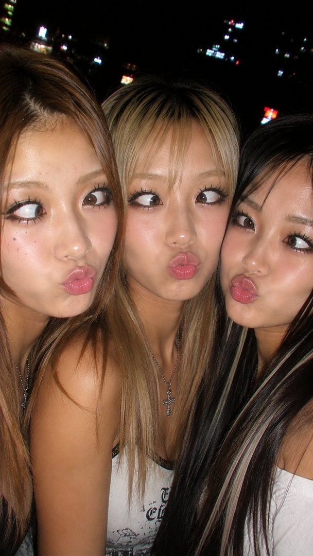 Three young East Asian women with pouty lips and playful expressions, posing for a selfie at night.