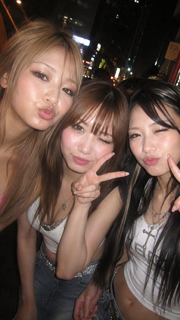 Three young East Asian women posing for a selfie at night in a city, making peace signs and pouting.