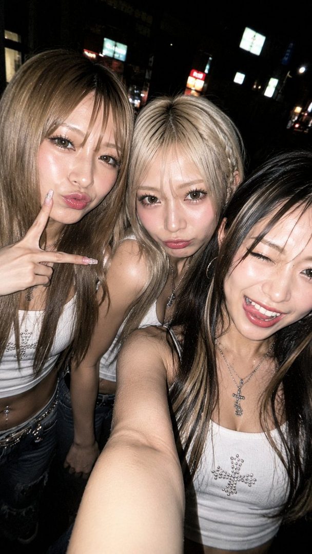 Three young women with long hair and fashionable outfits taking a selfie at night on a city street.