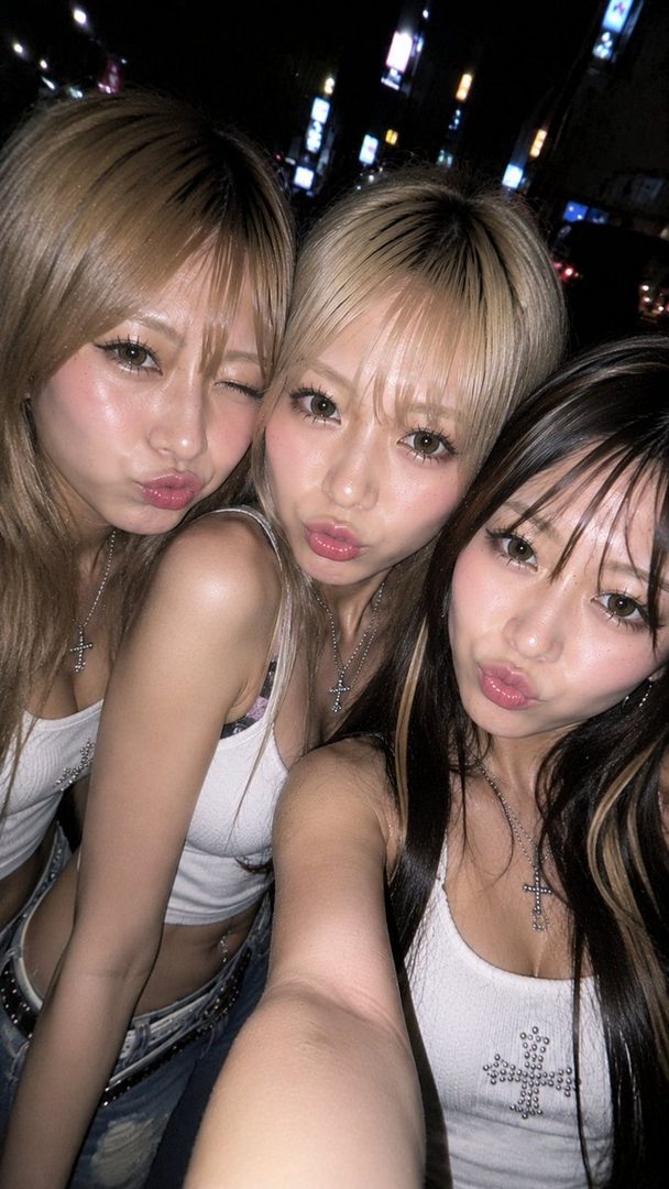 Three young women with blonde and brown hair taking a selfie at night, with city lights in the background.