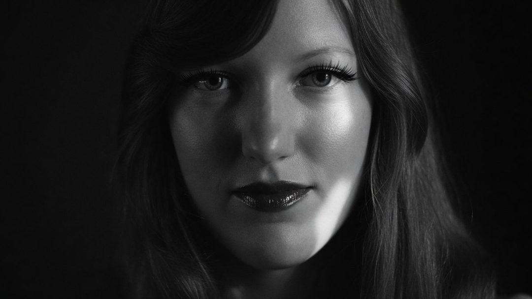 Close-up black and white portrait of a woman with long dark hair, dramatic lighting highlighting her face and dark lipstick.