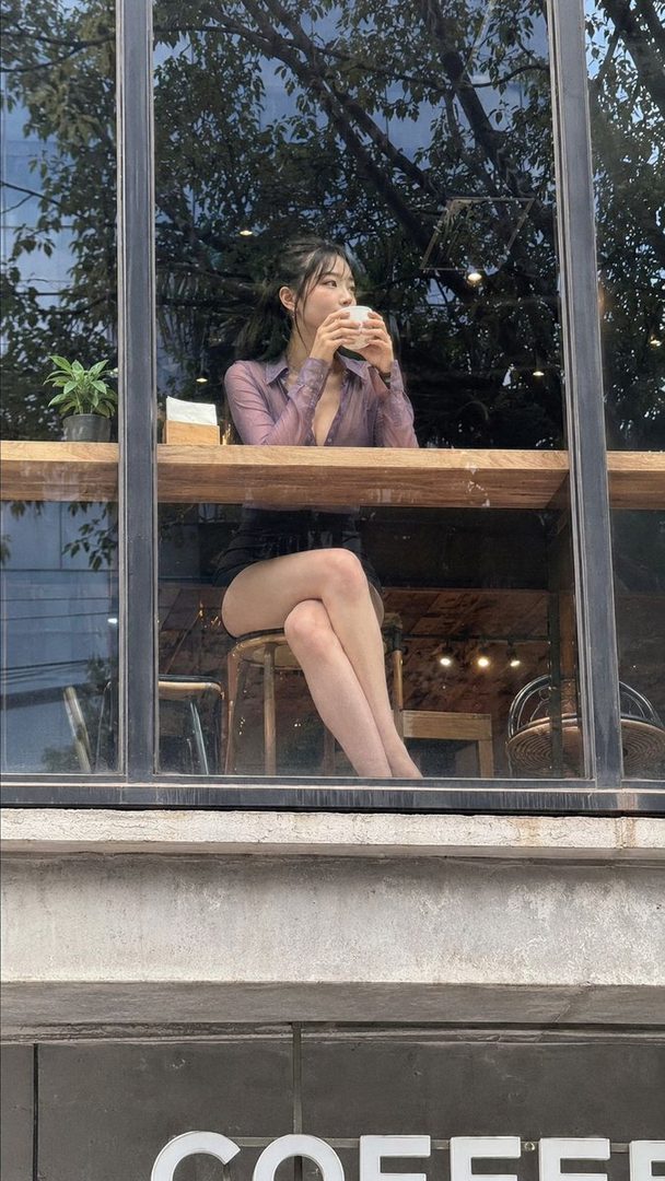 A young East Asian woman with long black hair tied in a ponytail, wearing a sheer purple blouse and dark shorts, sits at a cafe table by the window, holding a cup of coffee.