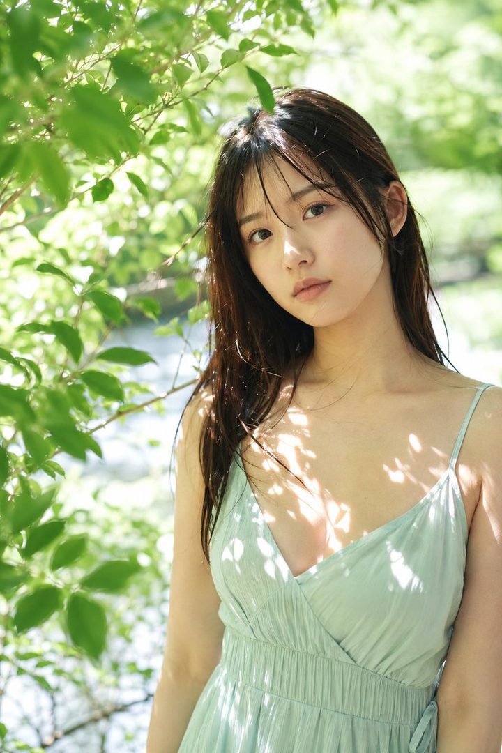 A young woman with long dark hair and a light green sundress stands outdoors, bathed in natural sunlight and dappled shadows from surrounding foliage.