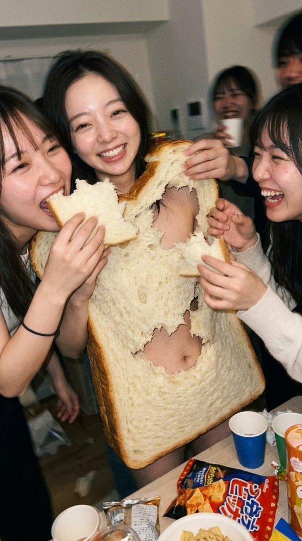 A group of young adults are playfully interacting with a large loaf of bread, with some taking bites and others posing.