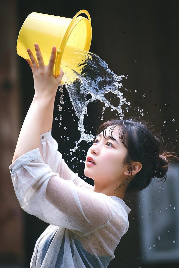 A young woman with her hair in a bun, wearing a sheer blouse, pours water from a yellow bucket, creating a splash.