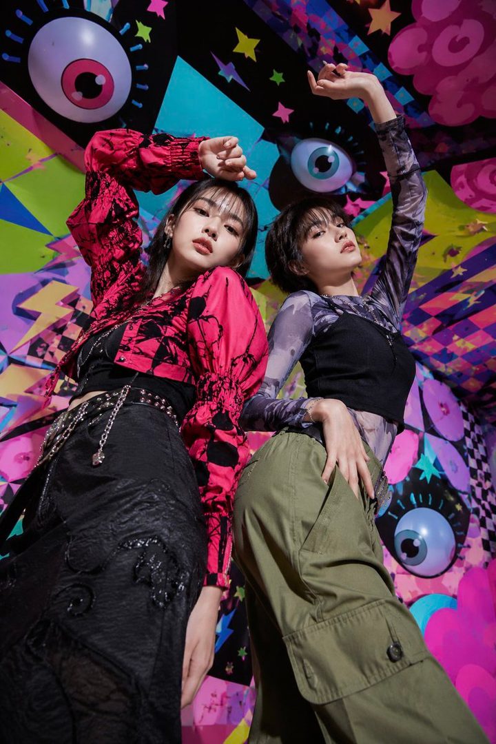 Two young East Asian women posing in front of a colorful, abstract background with large eye motifs.